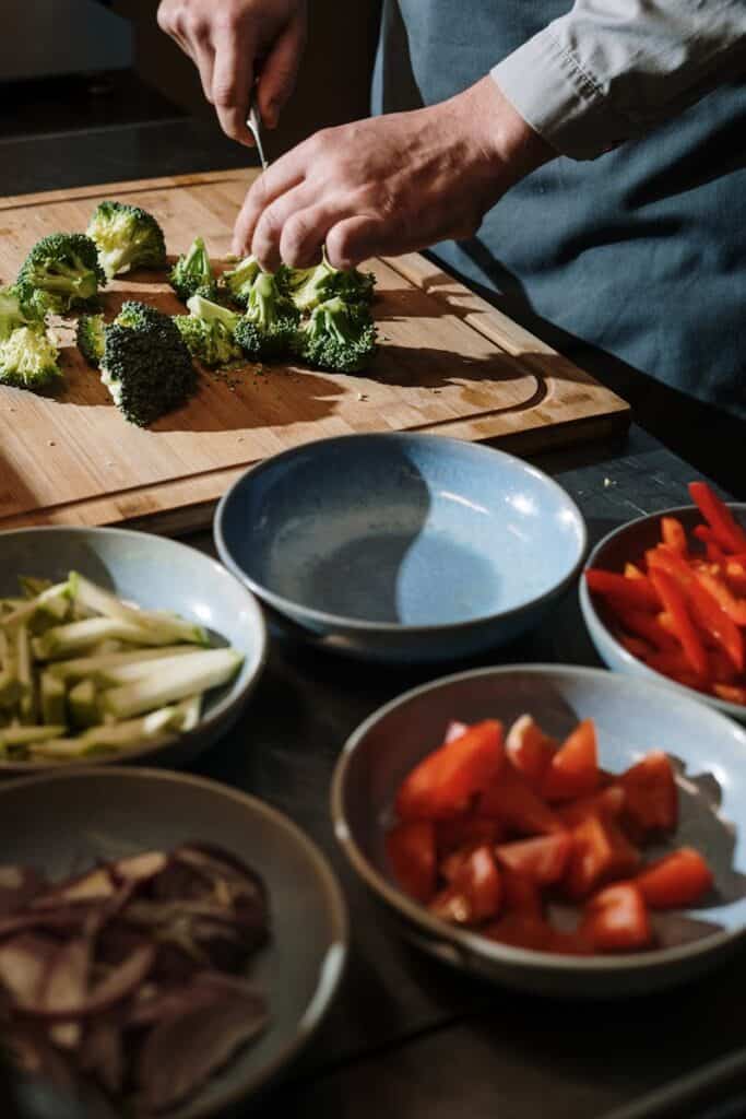 A chef chops broccoli on a cutting board surrounded by fresh vegetables in a kitchen setting.