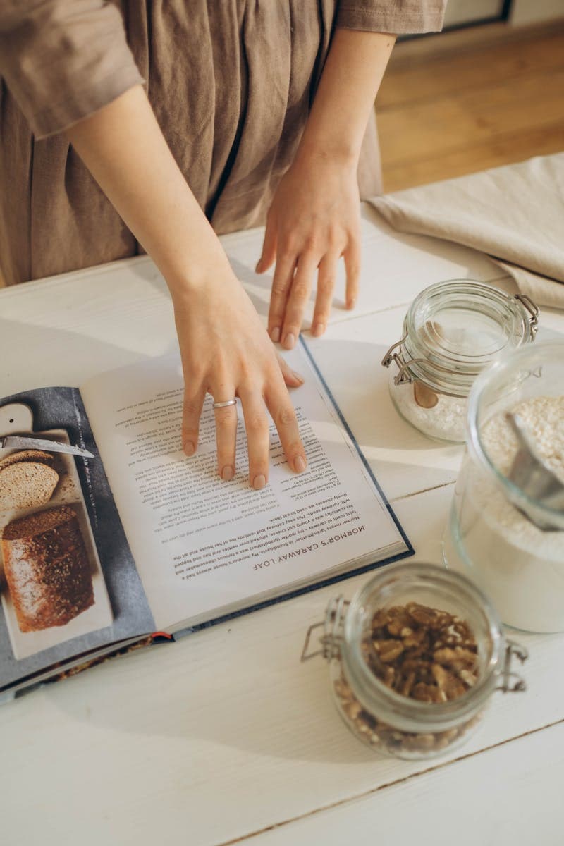 A person examines a cookbook surrounded by glass jars of ingredients on a kitchen table.