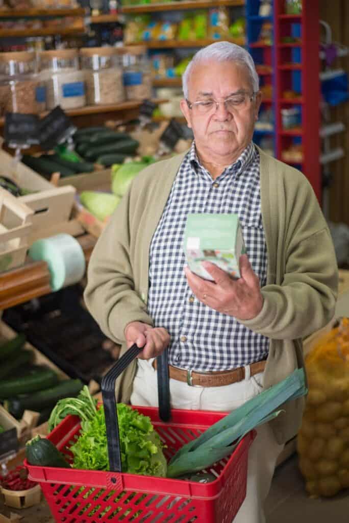 Elderly man holding a shopping basket in a grocery store, examining a product.